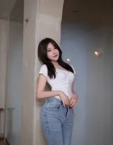 她坐在长椅上远眺，手轻抚发丝，牛仔裤与白衫在柔和光线下泛出质感，画面宁静致远