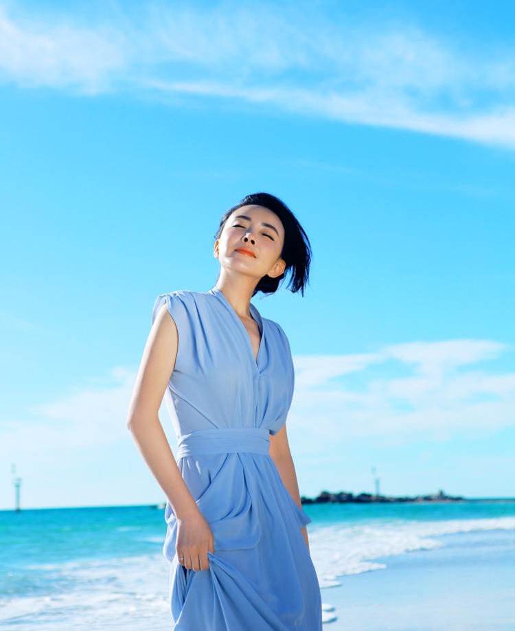 刘威葳身着飘逸白裙坐于沙滩礁石上，侧颜柔美，海天一色的背景将她衬托得如同海中精灵般纯净空灵
